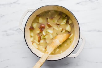 A white pot filled with creamy chowder, loaded with chunky potatoes, is shown on a marble surface, with a wooden spoon resting inside the pot.