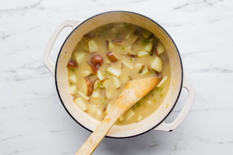 A white pot filled with creamy chowder, loaded with chunky potatoes, is shown on a marble surface, with a wooden spoon resting inside the pot.