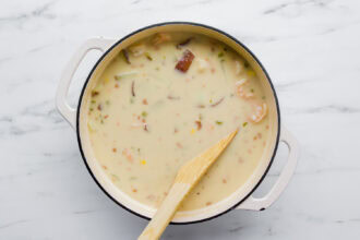 A white pot filled with creamy shrimp chowder, brimming with vegetables, sits on a marble surface with a wooden spatula resting in it.