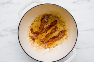 Three strips of cooked bacon in a white pot with rendered fat and browned bits, ready to add rich flavor as the base for a creamy chowder, set on a white marble surface.