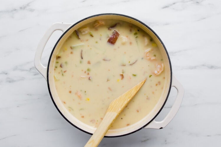 A white pot filled with creamy shrimp chowder, brimming with vegetables, sits on a marble surface with a wooden spatula resting in it.