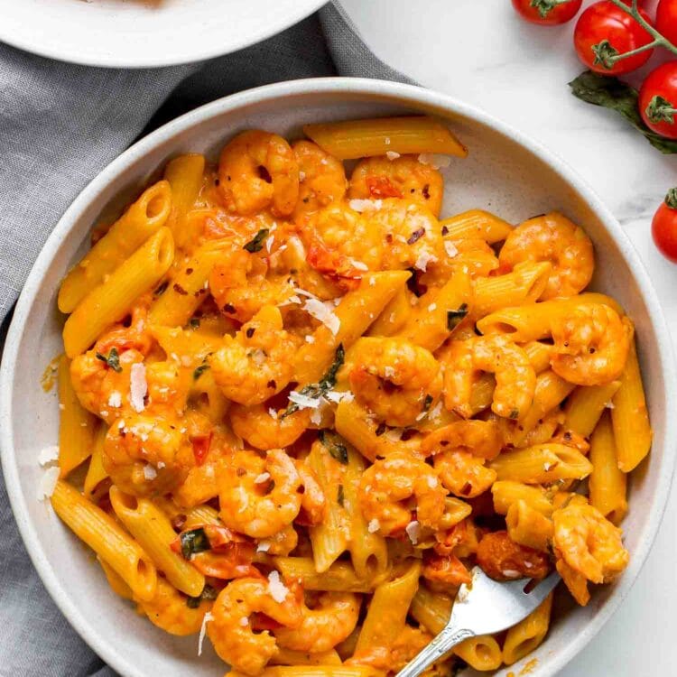A bowl of Creamy Tomato Shrimp Pasta garnished with herbs and grated cheese, served beside a fork and fresh cherry tomatoes.