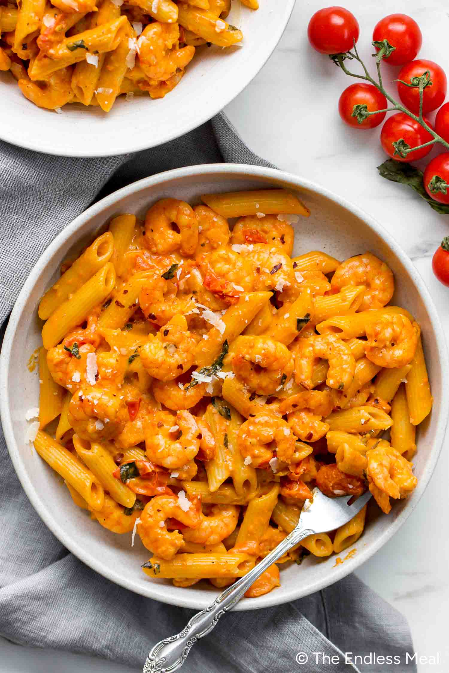 A bowl of Creamy Tomato Shrimp Pasta garnished with herbs and grated cheese, served beside a fork and fresh cherry tomatoes.