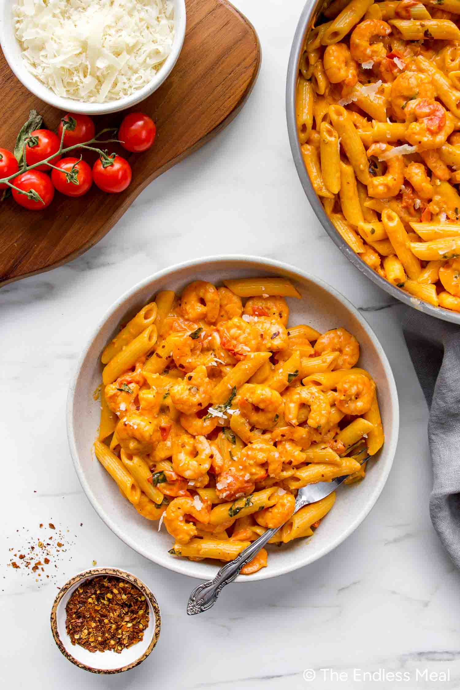 A bowl of Creamy Tomato Shrimp Pasta features penne with shrimp in a rich tomato sauce, topped with grated cheese, and served alongside bowls of cheese, cherry tomatoes, and red pepper flakes.