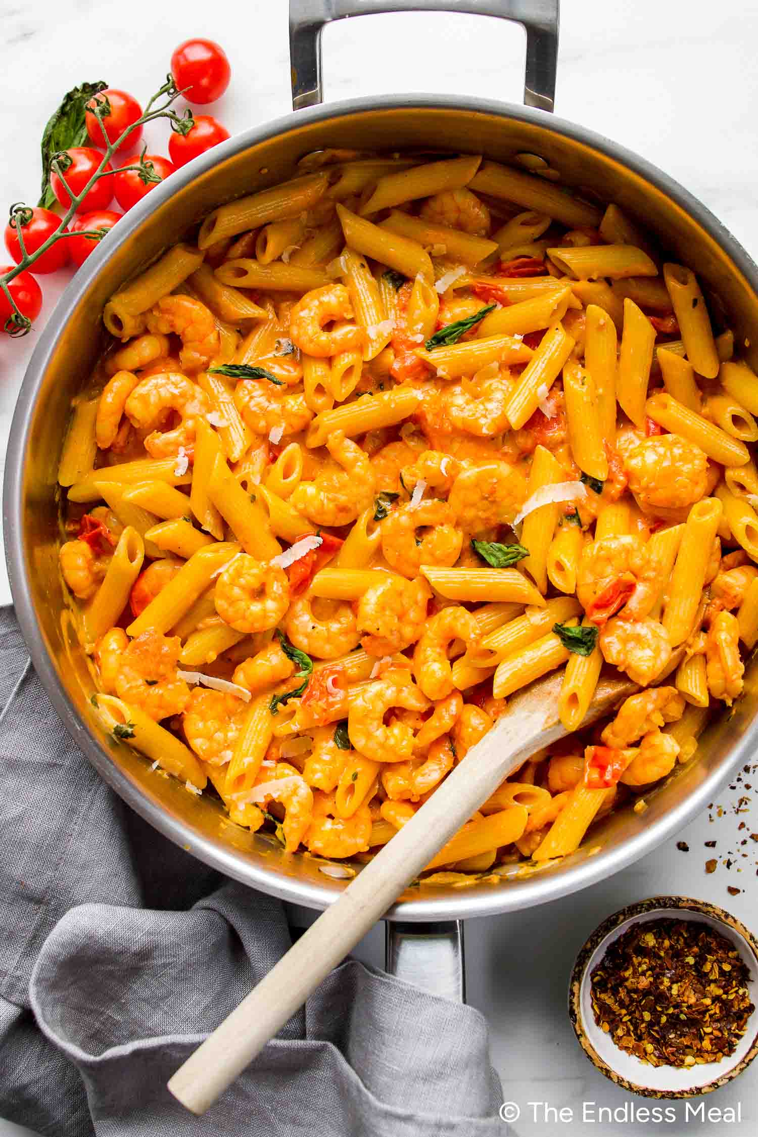 A pan of Creamy Tomato Shrimp Pasta with shrimp in a rich sauce, garnished with fresh basil, sits on a white surface beside cherry tomatoes and a small bowl of red pepper flakes.