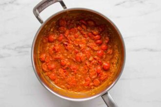 A stainless steel pan filled with cooked cherry tomatoes and sauce, perfect for tossing with creamy tomato shrimp pasta, sits on a white marble surface.