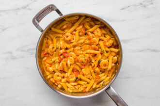 A stainless steel pan filled with Creamy Tomato Shrimp Pasta—penne, shrimp, cherry tomatoes, and a luscious orange sauce—all set on a marble countertop.