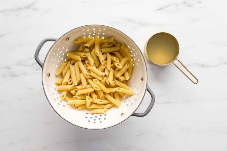 A colander filled with cooked penne pasta sits on a marble surface next to a metal measuring cup containing pasta water—perfect for preparing creamy tomato shrimp pasta.